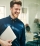 Young businessman laughing while walking in a modern office carrying a laptop with colleagues at work in the background